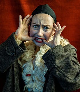 Alex Tatarsky stares at the camera, using their hands to pull their mouth into a smile. They wear bright red lipstick, a black cap, a feathered top and a green coat. They stand against a red fabric background under bright lighting.