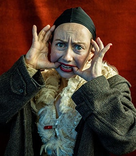 Alex Tatarsky stares at the camera, using their hands to pull their mouth into a smile. They wear bright red lipstick, a black cap, a feathered top and a green coat. They stand against a red fabric background under bright lighting.  