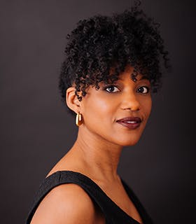 Tyler Thomas smiles over her right shoulder at the camera. Against a grey background, she wears a sleeveless black top and gold earrings.
