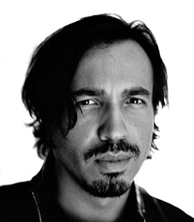A close up, black and white portrait of Rodrigo Valenzuela. He looks directly at the camera against a bright white background, wearing a black jacket.