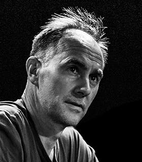 A close up, black and white portrait of David Watson. His face is turned away from the camera and he looks up and to the right, illuminated by an overhead light. He wears a grey T-shirt against a black background.