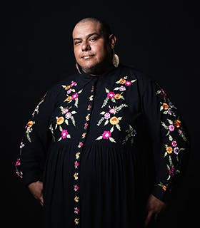 Dorian Wood stands smiling against a black background. Centered, she faces the camera with her hands in her pockets. She wears a black dress with embroidered flowers and leaf earrings.