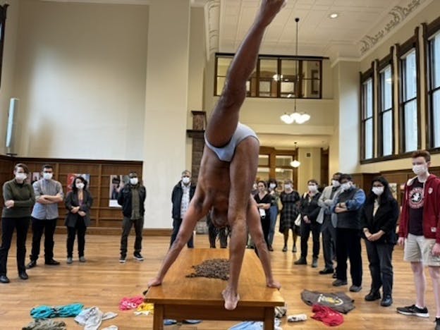 Javier Cardona Otero balances on a wooden table with two hands and one foot on the surface, while extending his other leg upward. He is wearing light gray briefs, and colorful clothing is scattered on the floor around the table. A pile of brown material is centered on the table. The performance takes place in a bright, spacious room with tall windows, wooden floors, and high ceilings, surrounded by an audience wearing masks.