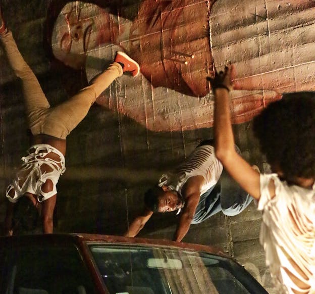 Javier Cardona Otero performs alongside two others in a darkly lit environment, on and around an abandoned car. They are positioned against a textured wall featuring a large mural of a person. One performer, in a distressed white shirt and jeans, leans on their hands, with their body angled downward, while another performer balances upside down on their hands, wearing beige pants and red sneakers. In the foreground, a third performer in torn white shirt raises an arm, partially out of focus.