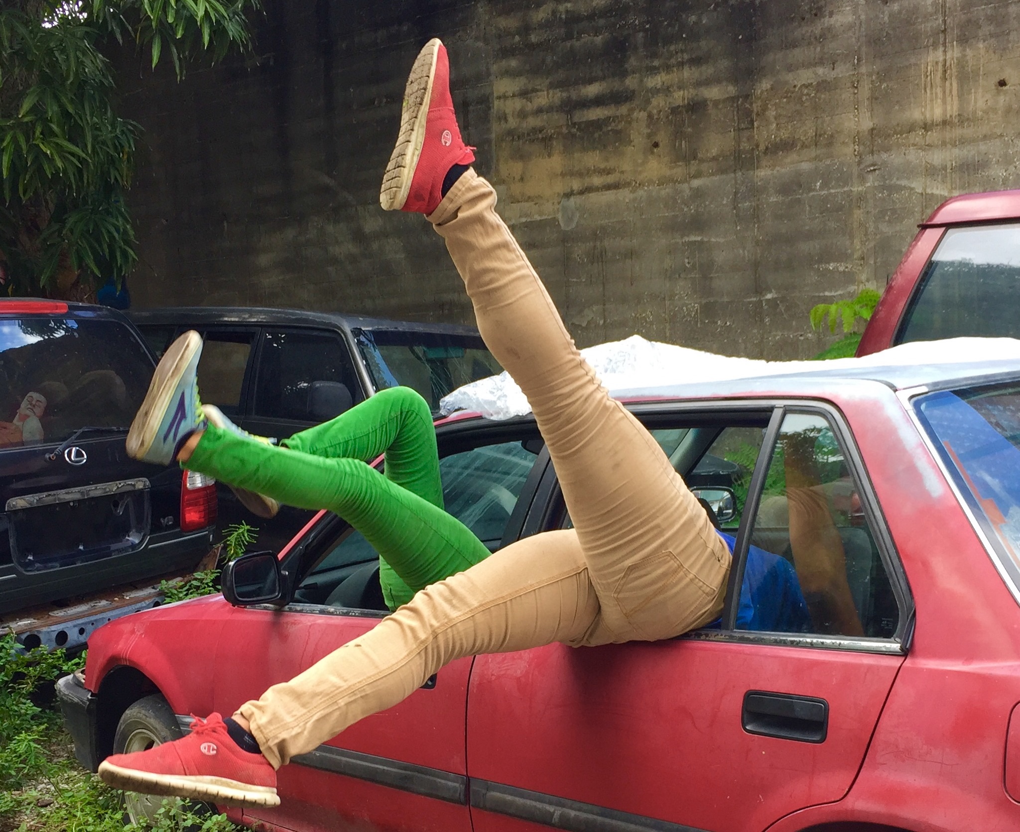 Two individuals extend their legs out of the windows of a red car, one wearing beige pants and red sneakers, the other wearing green pants and blue sneakers. The car is parked in a lot with other vehicles and surrounded by greenery, against the backdrop of a concrete wall.