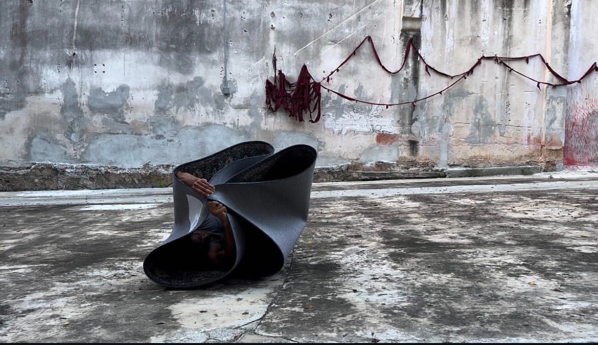 Javier Cardona Otero is partially visible inside a large, rolled piece of pliable material on a weathered concrete surface. Both of his hands extend out from the folds, gripping the edge of the material. The background features a worn, textured wall with peeling paint and draped red fabric strung along it.