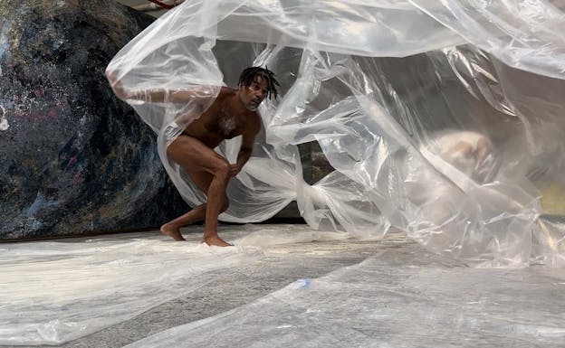 Javier Cardona Otero crouches in the foreground, partially wrapped in transparent plastic sheets. He is wearing black underwear and is positioned on a surface covered with plastic sheeting. Behind him, a large, textured, and multicolored spherical object is visible. Another figure is partially obscured by the plastic in the background.