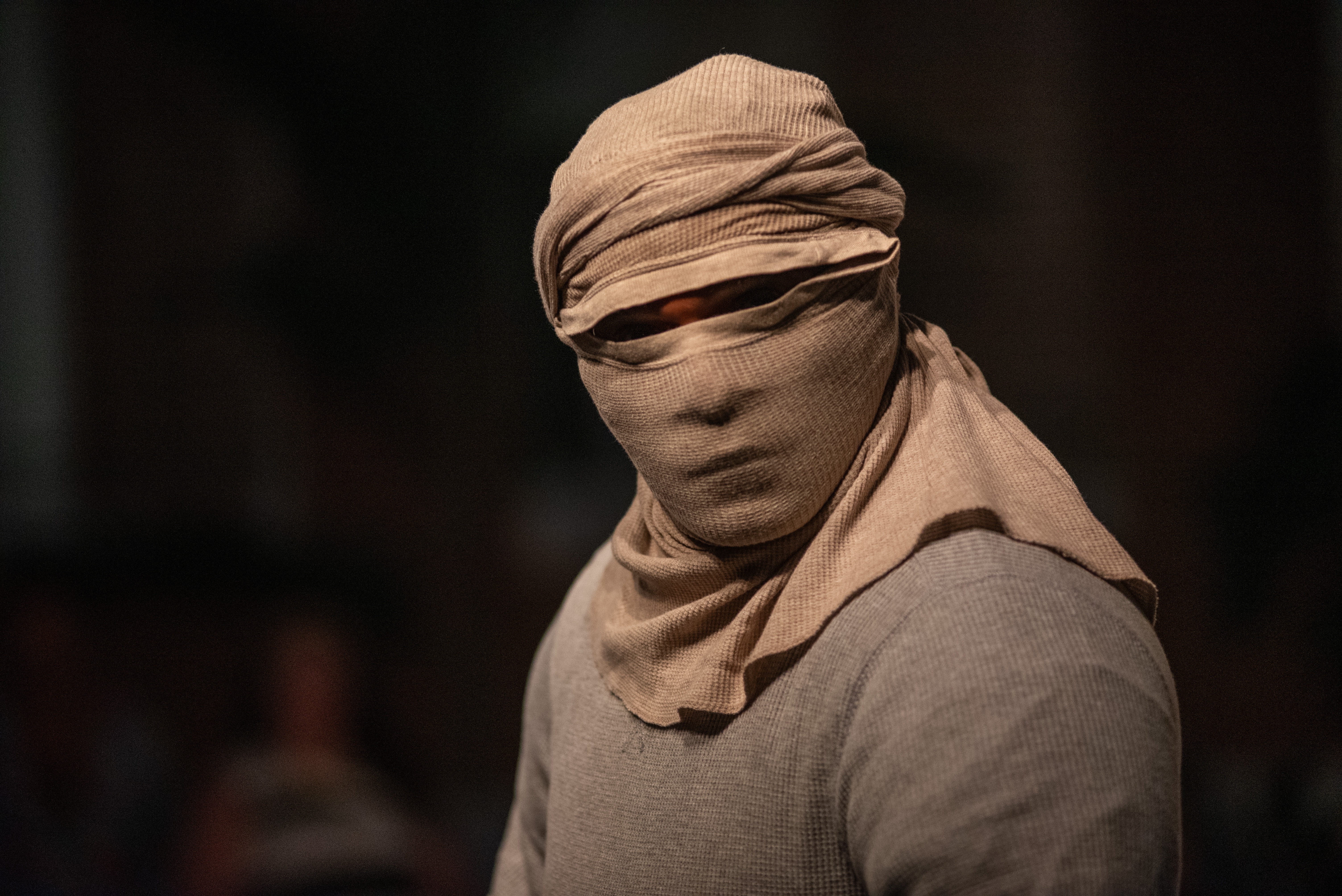 Javier Cardona Otero stands with his head and face fully wrapped in beige cloth, with only his eyes visible. He is wearing a gray shirt and is set against a dark, softly lit background.