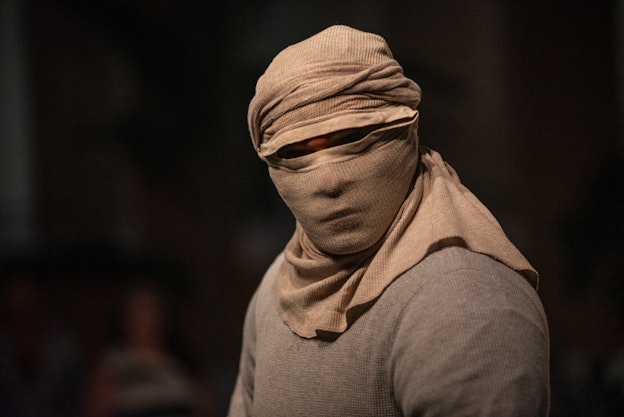 Javier Cardona Otero stands with his head and face fully wrapped in beige cloth, with only his eyes visible. He is wearing a gray shirt and is set against a dark, softly lit background.