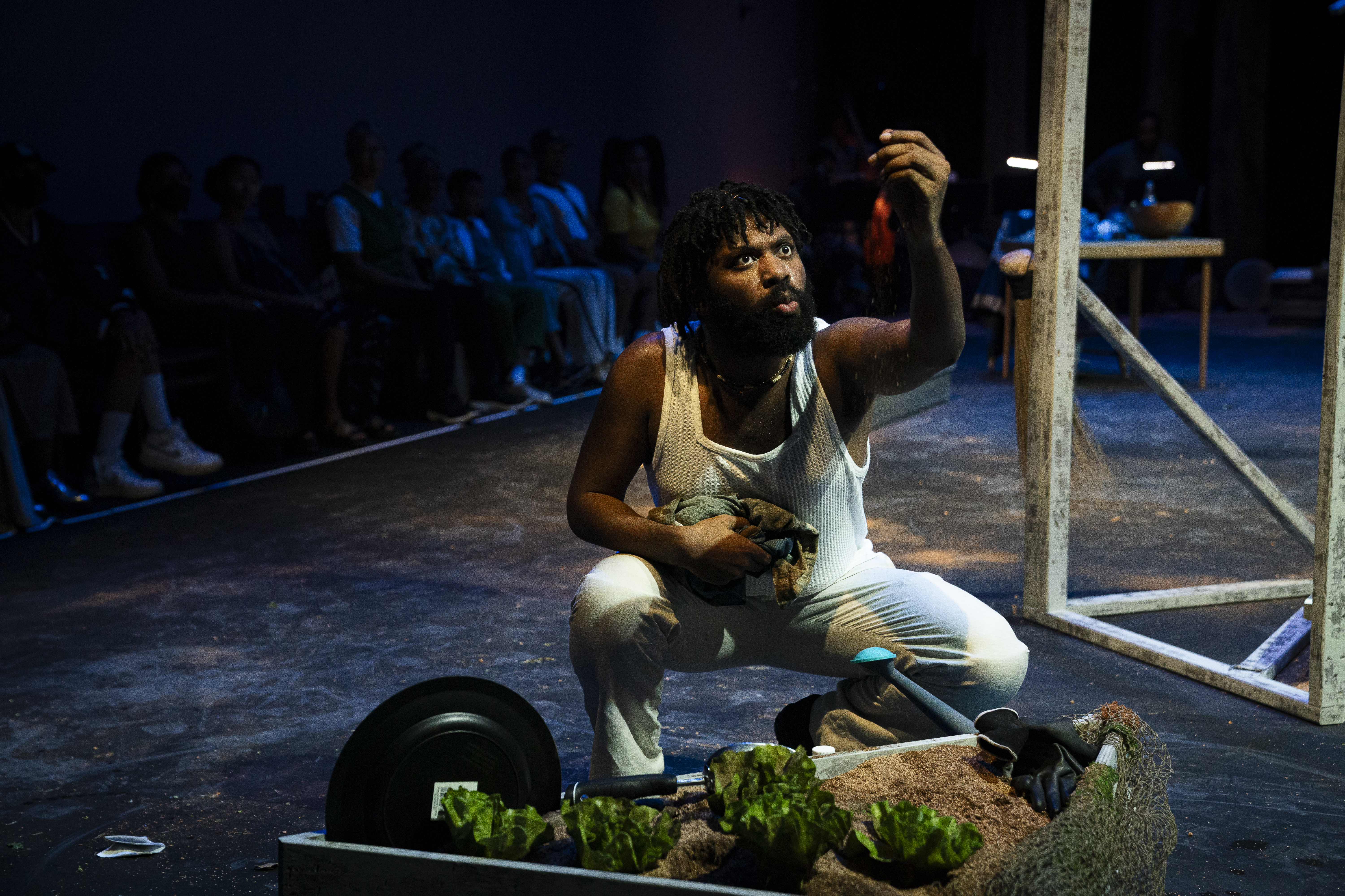 Trevor Hayes crouches on stage, holding a small piece of fabric in one hand and sprinkling sand from the other. They wear a textured mesh tank top and white pants, with gardening tools and a planter box filled with soil and leafy greens in the foreground. Elements of the stage installation are visible in the background, as are audience members seated in rows beyond the stage permiter.