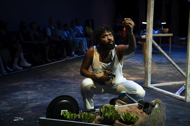 Trevor Hayes crouches on stage, holding a small piece of fabric in one hand and sprinkling sand from the other. They wear a textured mesh tank top and white pants, with gardening tools and a planter box filled with soil and leafy greens in the foreground. Elements of the stage installation are visible in the background, as are audience members seated in rows beyond the stage permiter.