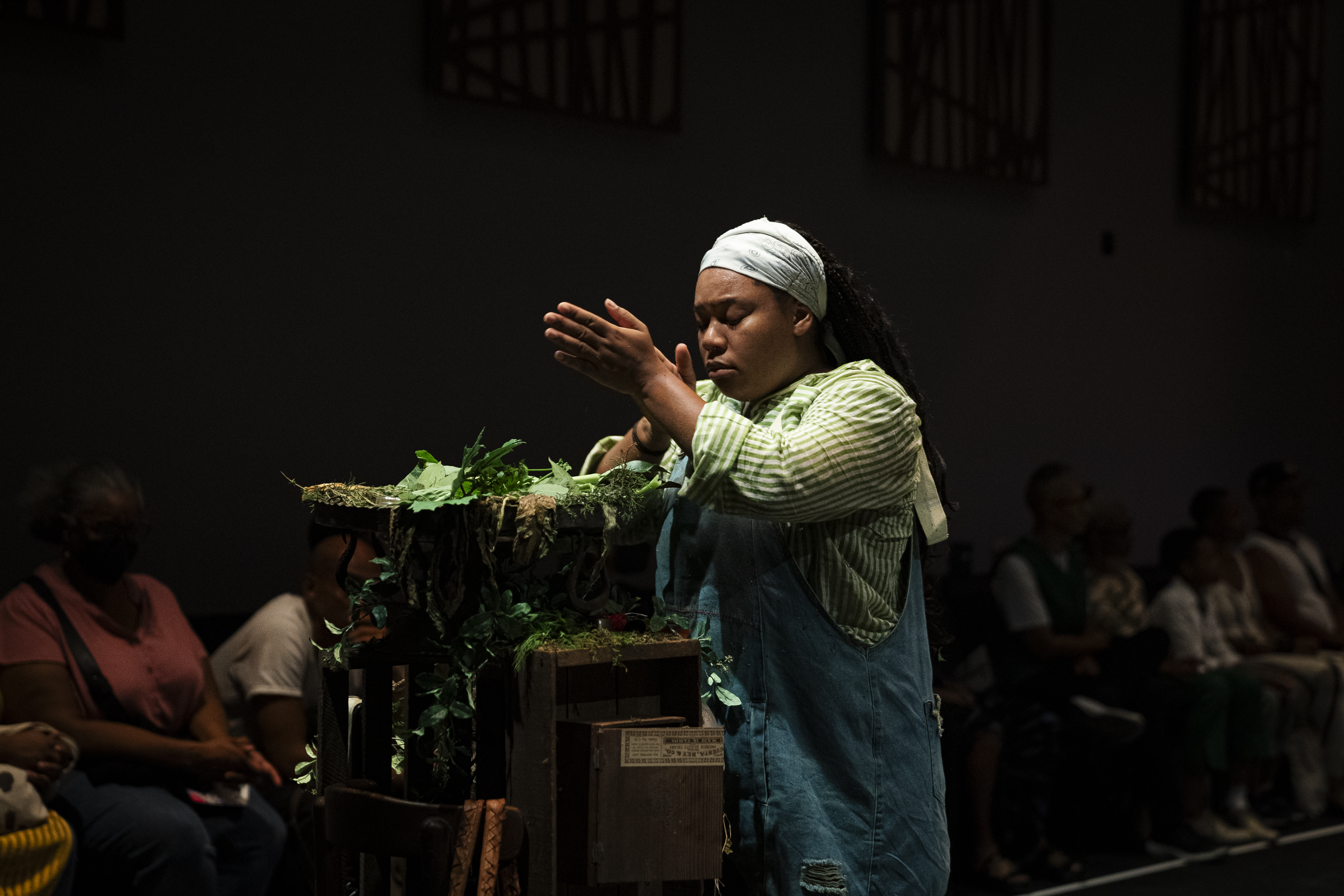 Sierra Leverett stands behind a structure covered in greenery and leaves, her hands raised and pressed together. She wears a light green striped shirt, blue denim overalls, and a light-colored bandana. The scene is dimly lit with an audience seated in the background.
