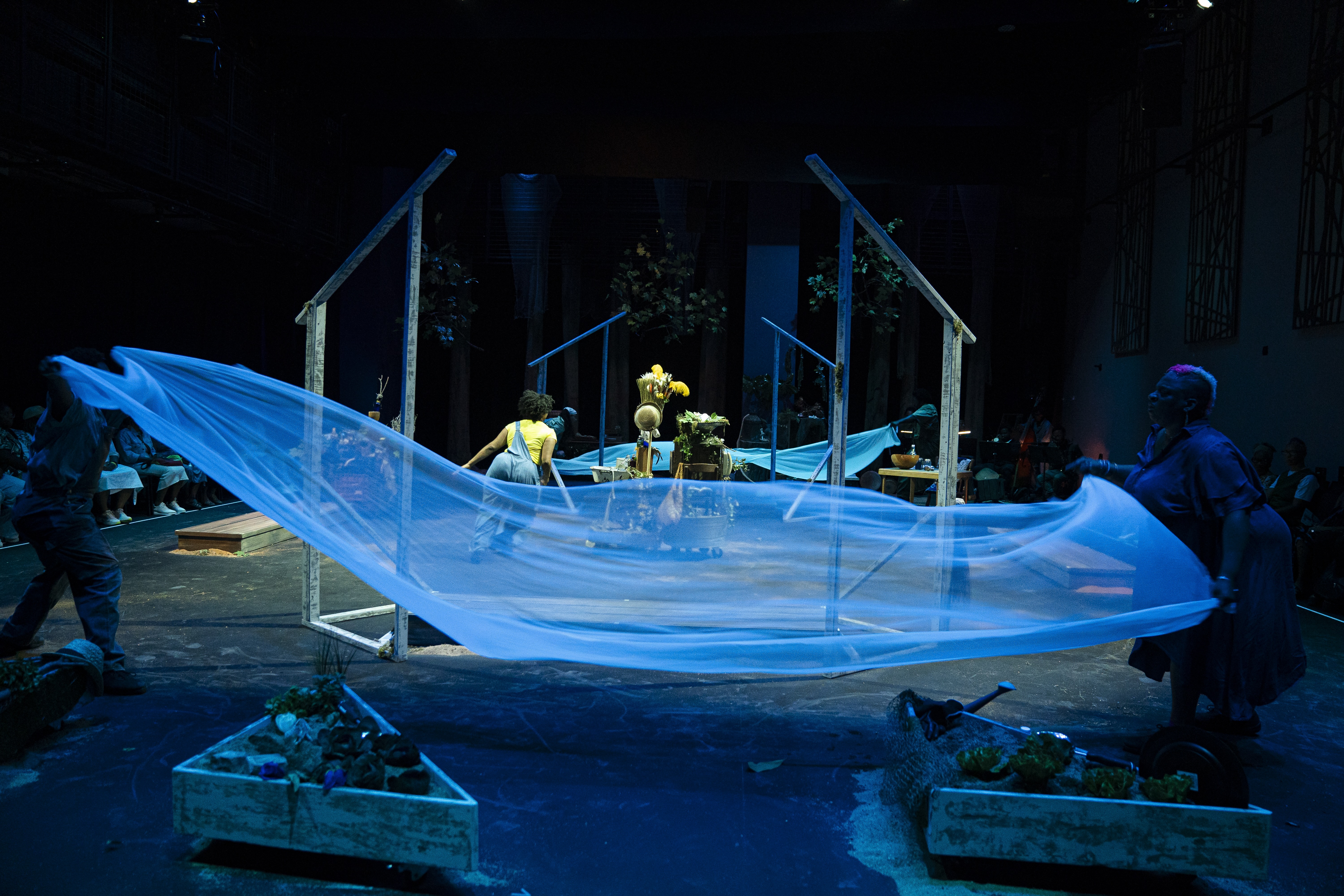 Sierra Leverett, Sheshe Dance, Fletcher Laws, Trevor Hayes, Rayna Johnson, and Zenni Corbin interact with a stage installation comprised of flowing blue fabric stretched between wooden frames. The stage contains plants and other natural elements arranged across the space, and is illuminated with blue lighting. In the background, greenery and additional props are visible. The audience is seated on either side of the performance area.