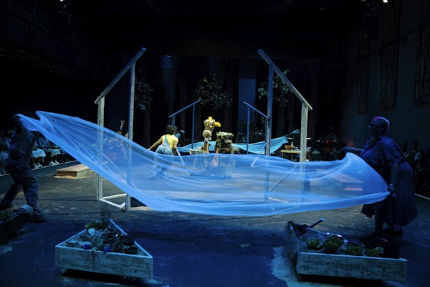 Sierra Leverett, Sheshe Dance, Fletcher Laws, Trevor Hayes, Rayna Johnson, and Zenni Corbin interact with a stage installation comprised of flowing blue fabric stretched between wooden frames. The stage contains plants and other natural elements arranged across the space, and is illuminated with blue lighting. In the background, greenery and additional props are visible. The audience is seated on either side of the performance area.