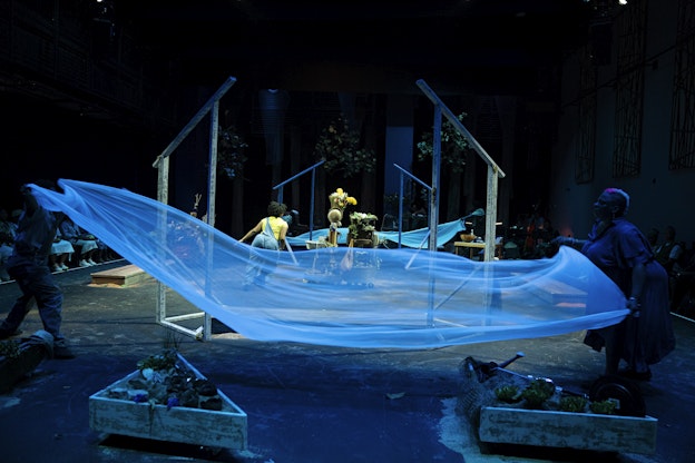 Sierra Leverett, Sheshe Dance, Fletcher Laws, Trevor Hayes, Rayna Johnson, and Zenni Corbin interact with a stage installation comprised of flowing blue fabric stretched between wooden frames. The stage contains plants and other natural elements arranged across the space, and is illuminated with blue lighting. In the background, greenery and additional props are visible. The audience is seated on either side of the performance area.