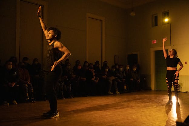 Zenni Corbin and Ebony Webster stand a distance apart on a dimly lit stage, each raising one arm into the air. Zenni Corbin, on the left, wears a sleeveless black mesh top and black pants, with one hand placed on their hip. Ebony Webster, on the right, stands towards the back of the stage and wears a black cropped shirt and black pants with cutouts along the legs. The audience is seated in the background, with many wearing masks.