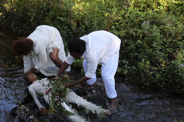 Ebony Webster, Jude Evans, and Fletcher Laws are in a shallow stream surrounded by lush greenery. One performer lies in the water with their eyes closed, while the other two stand to their side. The downstream performer holds a bunch of flowers and foliage over the person lying in the stream, and the upstream performer gently places a hand on their forehead. All three wear white outfits.