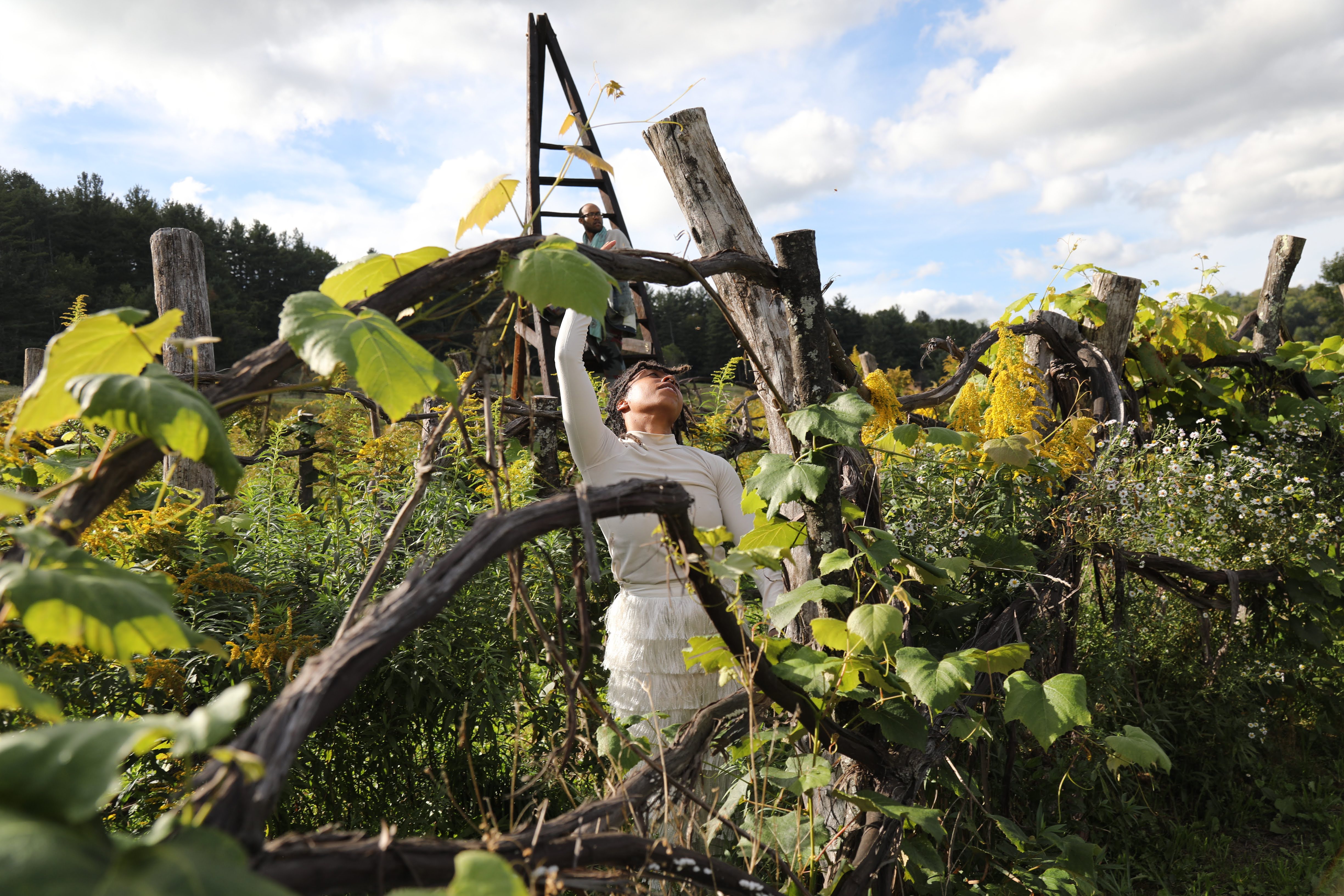 Ebony Webster stands among overgrown greenery and wooden branches in an outdoor setting. Wearing a white, long-sleeved top and a textured white skirt, they extend one arm upward, gazing toward the sky. In the background, a large wooden structure and another figure are partially visible, with a backdrop of trees and a blue sky with scattered clouds.