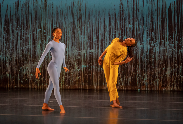 Shayla-Vie Jenkins and Juliette Jones stand on a stage in front of a backdrop featuring blue vertical textured patterns. Juliette Jones, dressed in a light blue body suit, stands upright with arms extended at their sides, while Shayla-Vie Jenkins, dressed in yellow, stands on the right leaning to the side with her head tilted and arms curved.