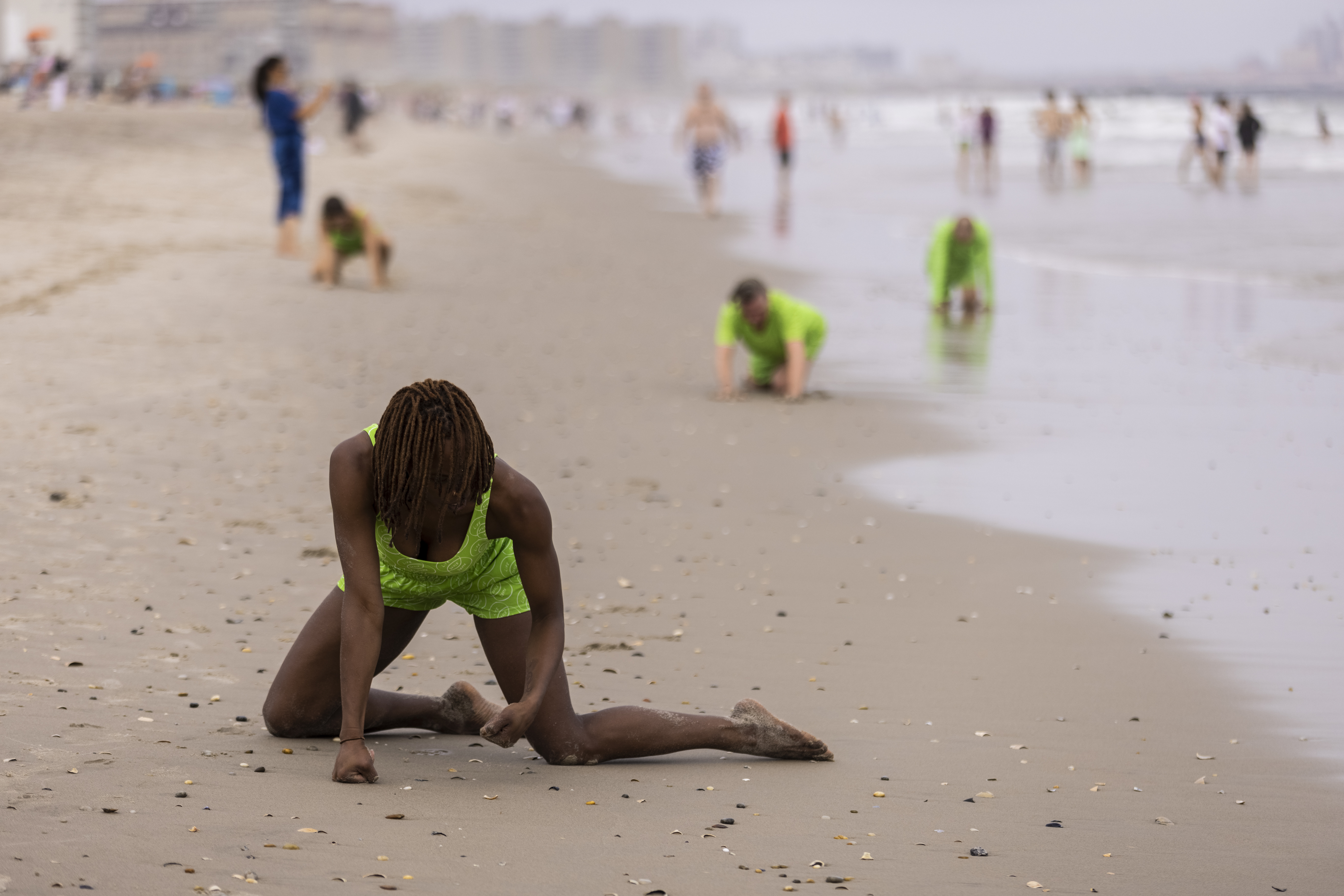 Shayla-Vie Jenkins kneels on a beach in a neon green outfit, pressing her fists into the ground as she extends her legs sideways. The sky is overcast and, in the background, additional performers dressed in similar neon green outfits crawl across the sand. Beachgoers are seen in the distance walking along the shoreline.