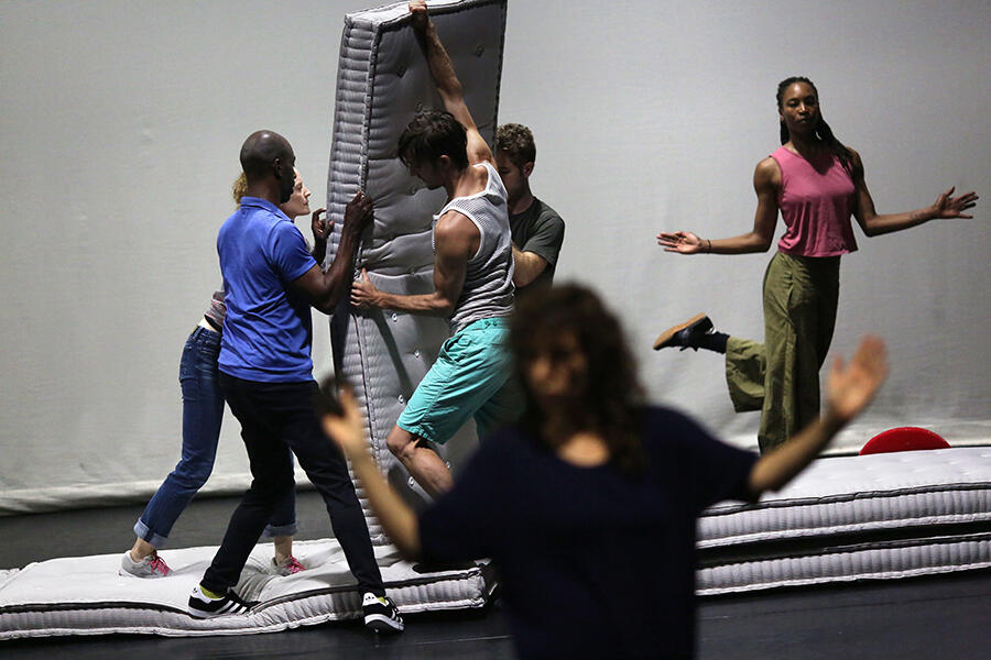 Four performers are shown bracing a mattress on all sides while Shayla-Vie Jenkins stands on one foot in the background with both arms extended outward. The stage upon which they stand is comprised of several stacked mattresses. The performance space is brightly lit and another performer stands in the foreground of the shot, appearing blurry.