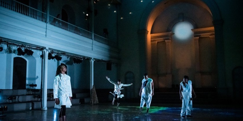 Four performers dressed in white clothing move across an open stage that is dimly lit, each illuminated by blue and orange toned lighting. The space features high ceilings, an archway, and ornate architectural elements. One of the performers standing in the far back of the stage is captured mid-motion with their arms extended outwards.