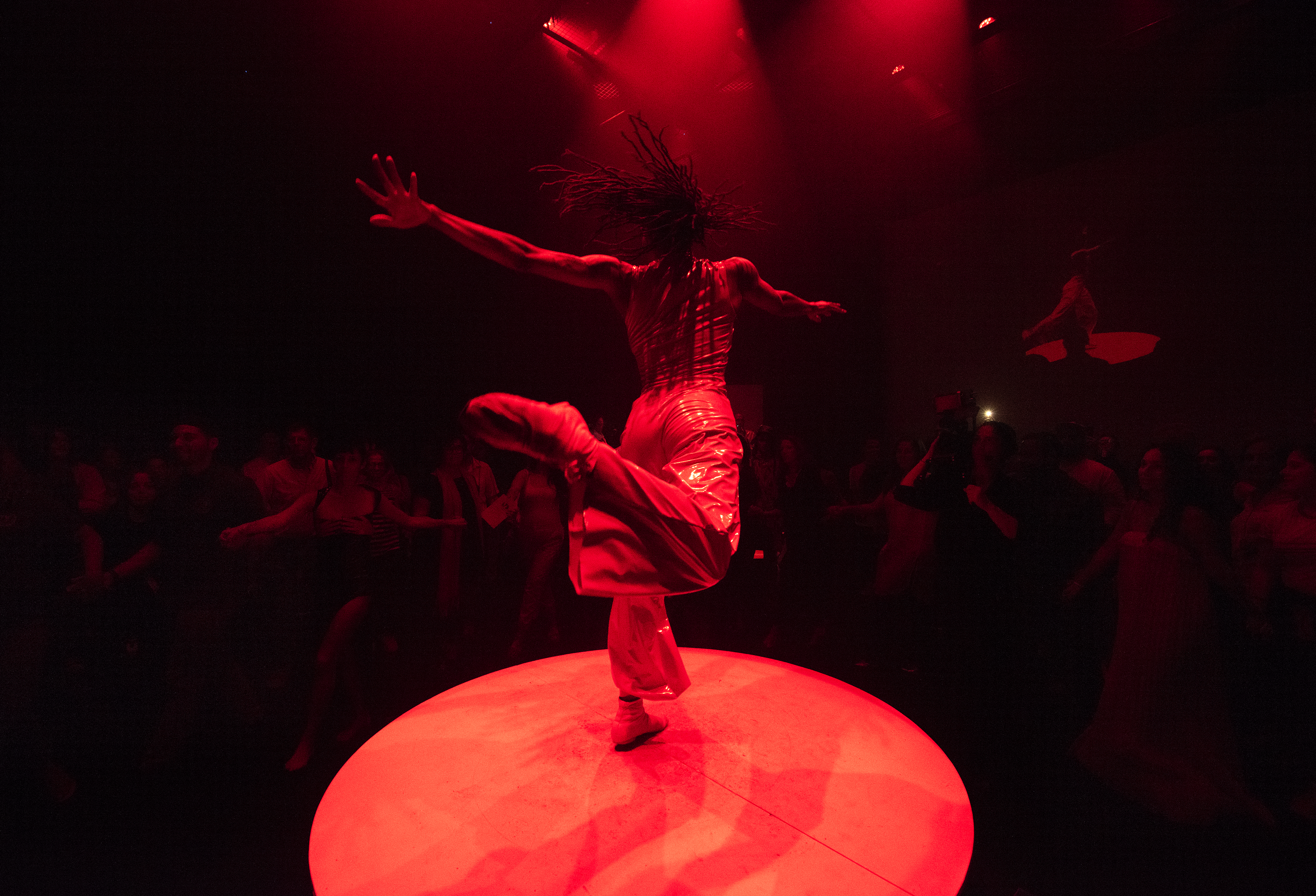 Shayla-Vie Jenkins faces the audience as she performs on a raised circular platform under intense red lighting. She is captured in motion with her arms are outstretched and one leg bent backwards.