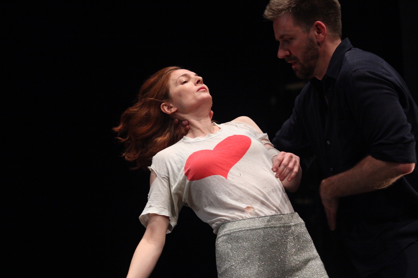Jodi Melnick and Jon Kinzel are dancing together, with Melnick leaning backward while Kinzel supports her with one hand behind her neck. Melnick is wearing glittery grey pants and a tattered white shirt with a red heart graphic and Kinzel is wearing a half sleeve dark blue shirt. They are positioned in front of a dark stage backdrop.