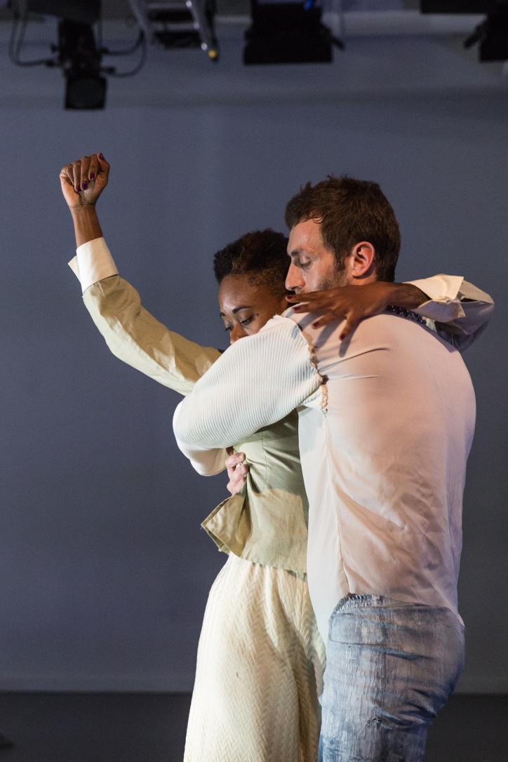 Omagbitse Omagbemi and Simon Courchel are captured in an embrace, with Omagbemi's arm raised in a fist and Courchel's arms wrapped around Omagbemi. Omagbemi wears a light green long sleeve shirt and skirt, while Courcel wears light-blue pants and a white long sleeve shirt. They are framed against a neutral gray background with soft lighting.