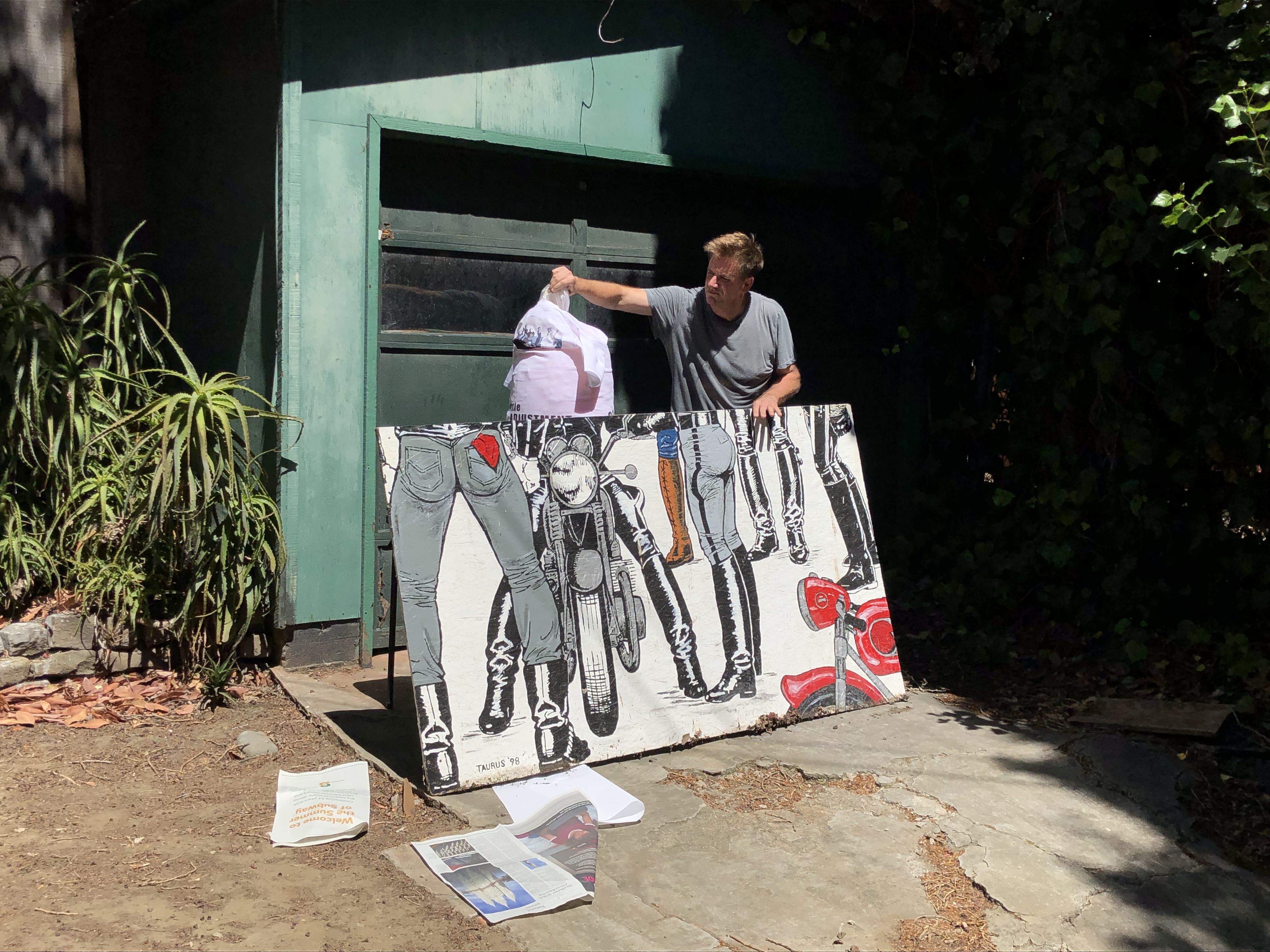 Jon Kinzel is outdoors standing behind a painted rectangular canvas tilted on its side and propped up against a column-like sculpture in front of a green garage door. The canvas features ink illustrations depicting legs, leather boots, and motorcycles. There is foliage visible and scattered newspapers are seen on the ground.
