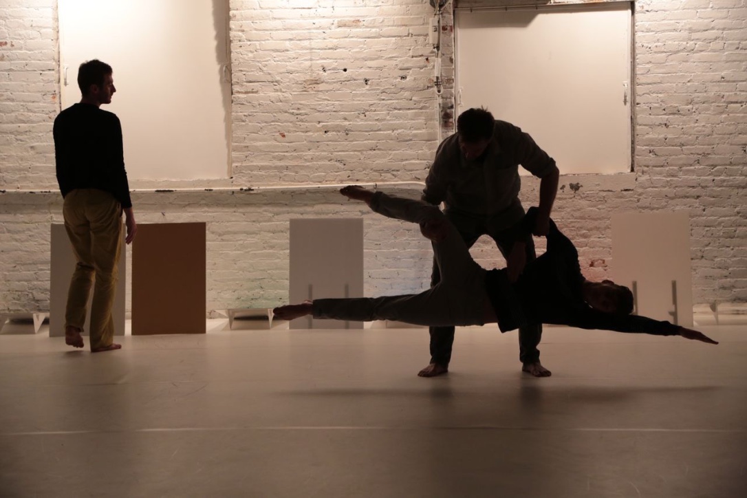 Simon Courchel, Stuart Shugg, and Jon Kinzel perform in a dimly lit room with exposed brick walls. The three performers appear as silhouettes, with Kinzel carrying one of the performers, who is in a horizontal pose with their arms outstretched. Minimal rectangular stage props are seen in the space.