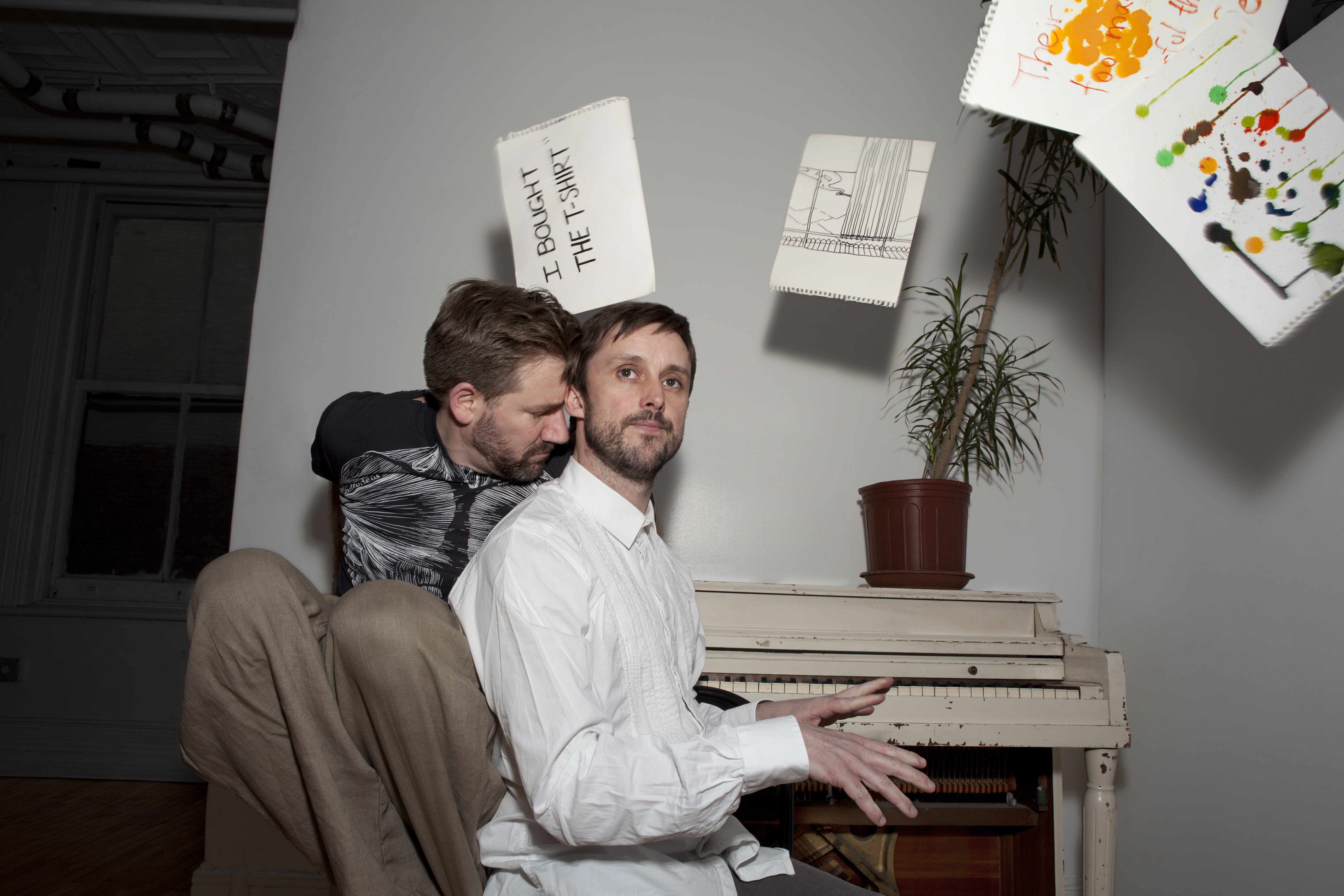 Philip Connaughton and Jon Kinzel are seated at a piano in a room with floating sheets of paper around them. Kinzel, wearing a black tee, leans closely into Connaughton, who is poised to play the keys, wearing a white button-down shirt.