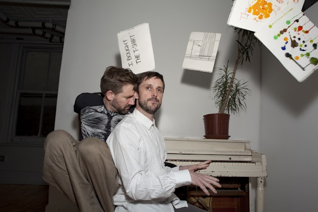 Philip Connaughton and Jon Kinzel are seated at a piano in a room with floating sheets of paper around them. Kinzel, wearing a black tee, leans closely into Connaughton, who is poised to play the keys, wearing a white button-down shirt.