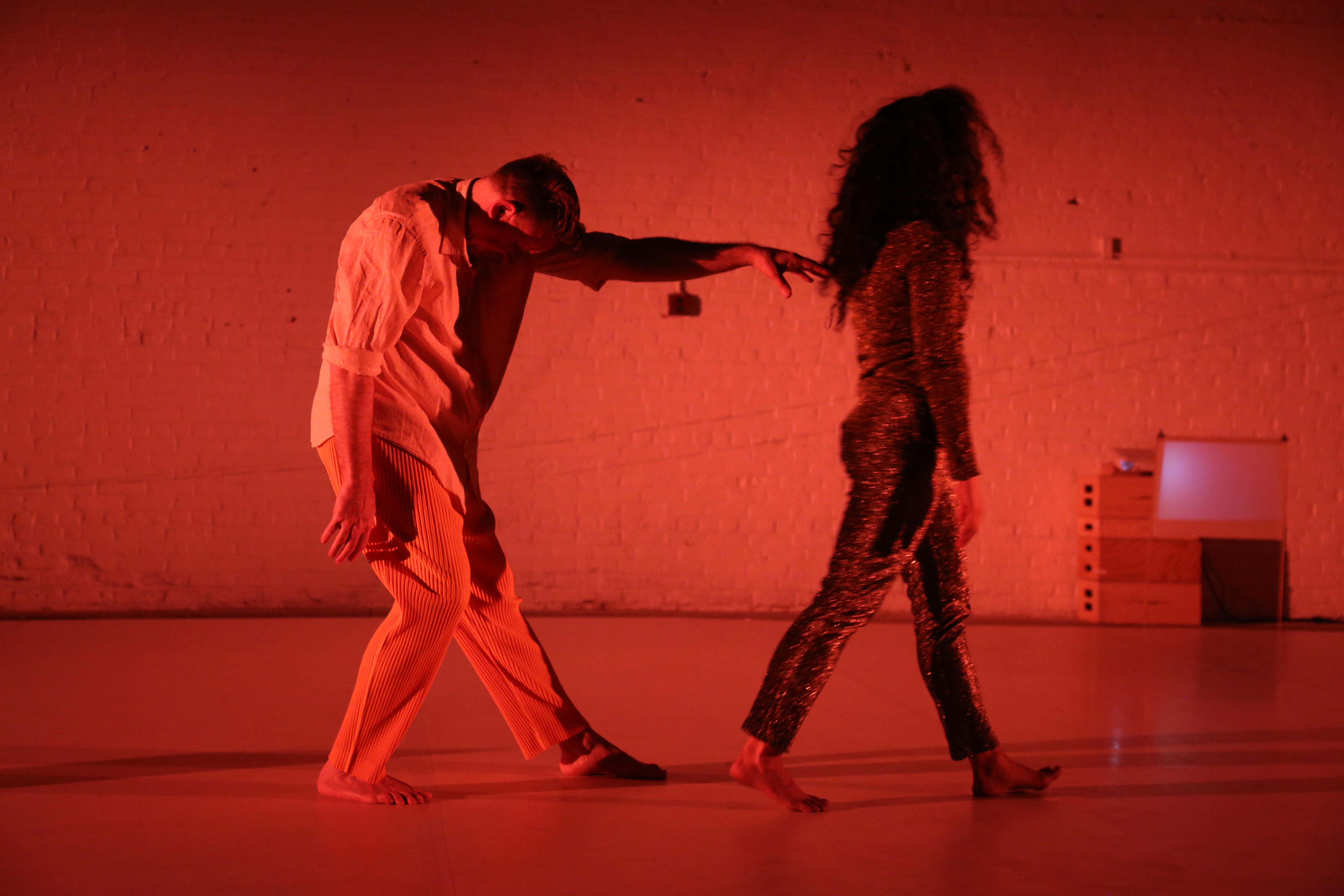 Priscilla Marrero and Jon Kinzel perform on an empty stage in front of a brick wall completely covered in red lighting. Marrero walks away in a shimmering, textured outfit while Kinzel, in a loose shirt and striped pants, leans forward with one arm stretched out towards Marrero. Their movements are captured mid-step.