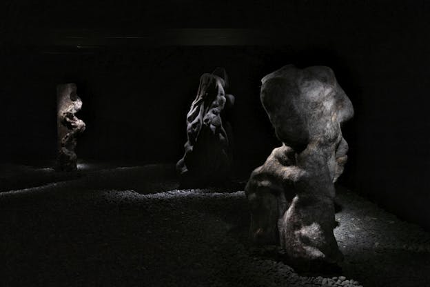 Three organic, abstract felted wool sculptures stand in a dimly lit gallery on a floor covered in gravel. Spotlights highlight their textured surfaces, revealing folds, crevices, and shadowy contours.