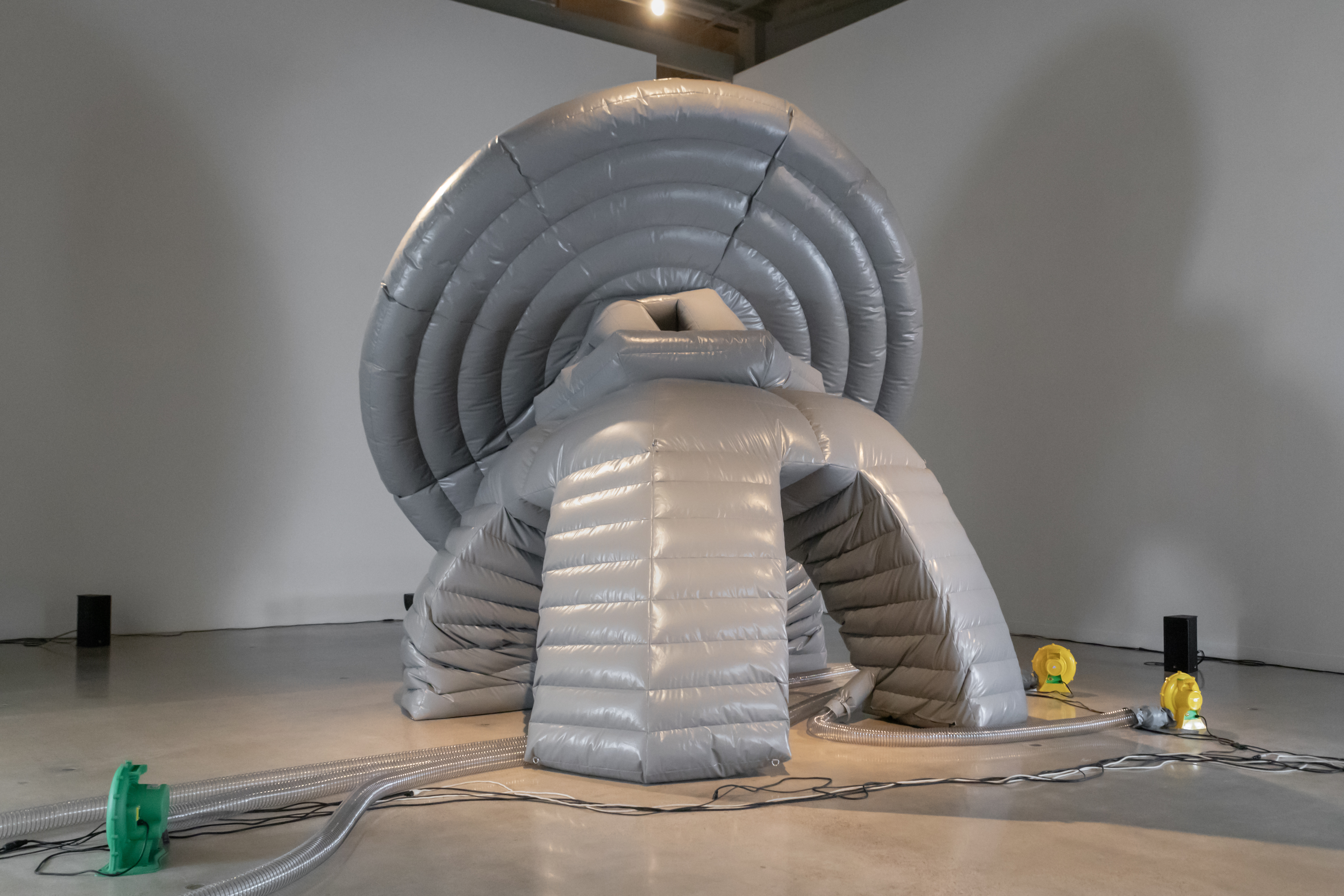 A large, inflatable grey structure with ribbed, circular sections and arched openings occupies the center of a gallery. Tubes and electrical cords extend from the base to nearby equipment. The sculpture stands atop polished concrete floors and is surrounded by white walls.