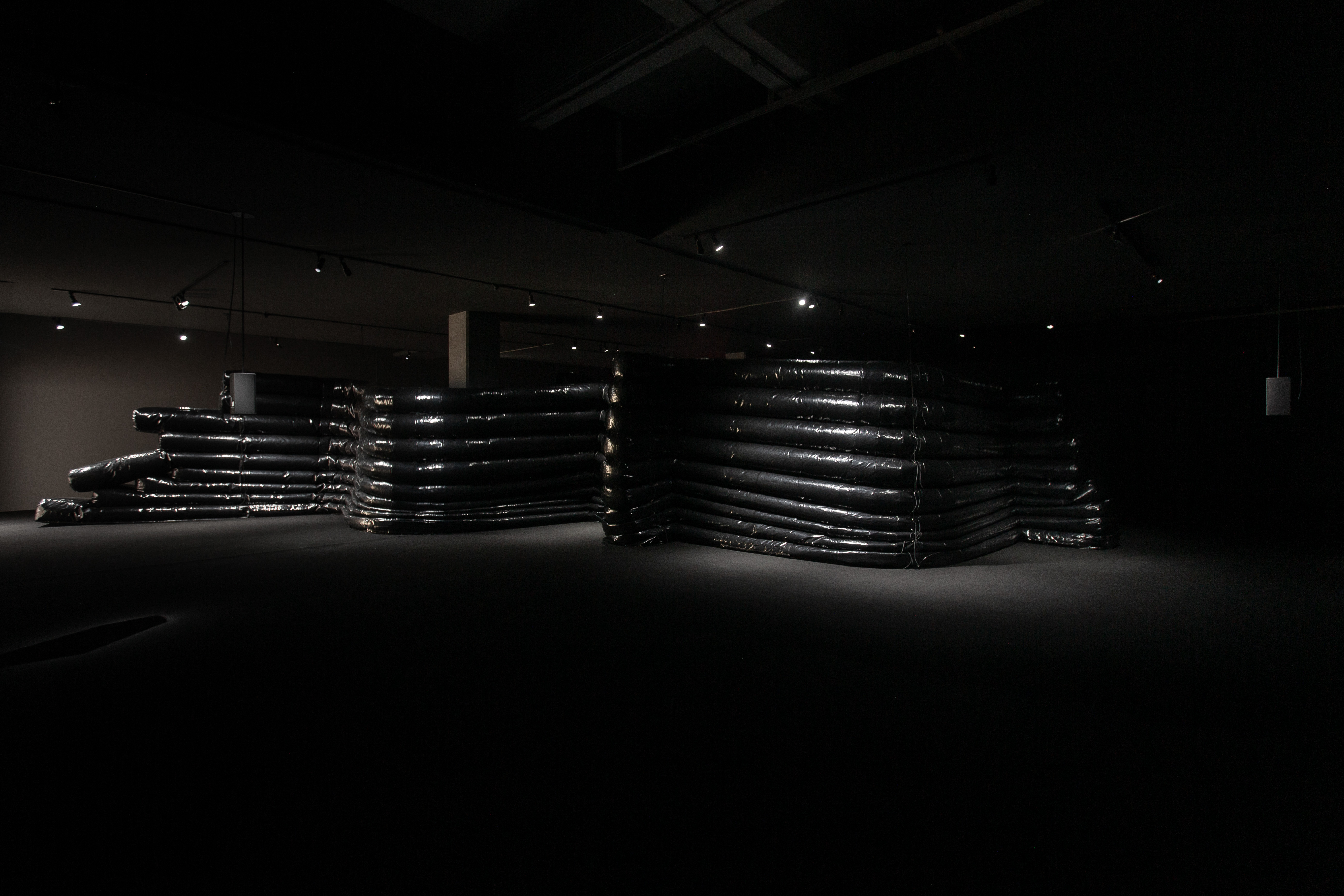 A darkened gallery space filled with stacked black inflatable tubes arranged into large, labyrinth-like forms. Subtle lighting highlights the glossy surface of the tubes, while the surrounding area remains dim.