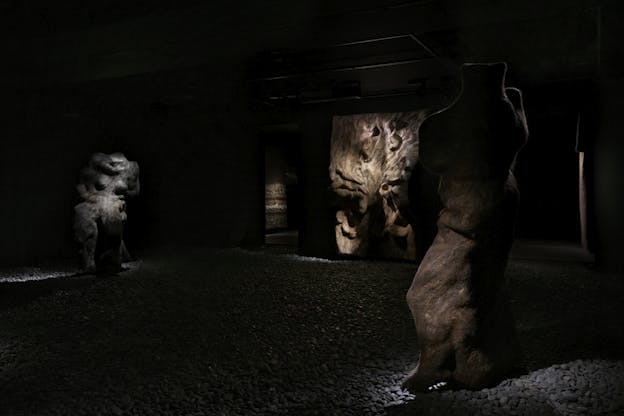 A dark gallery space featuring three large, felted wool sculptures on a gravel-covered floor. Soft lighting casts dramatic shadows that highlight their layered, uneven surfaces. In the background, a wall-mounted piece with intricate folds is faintly illuminated.