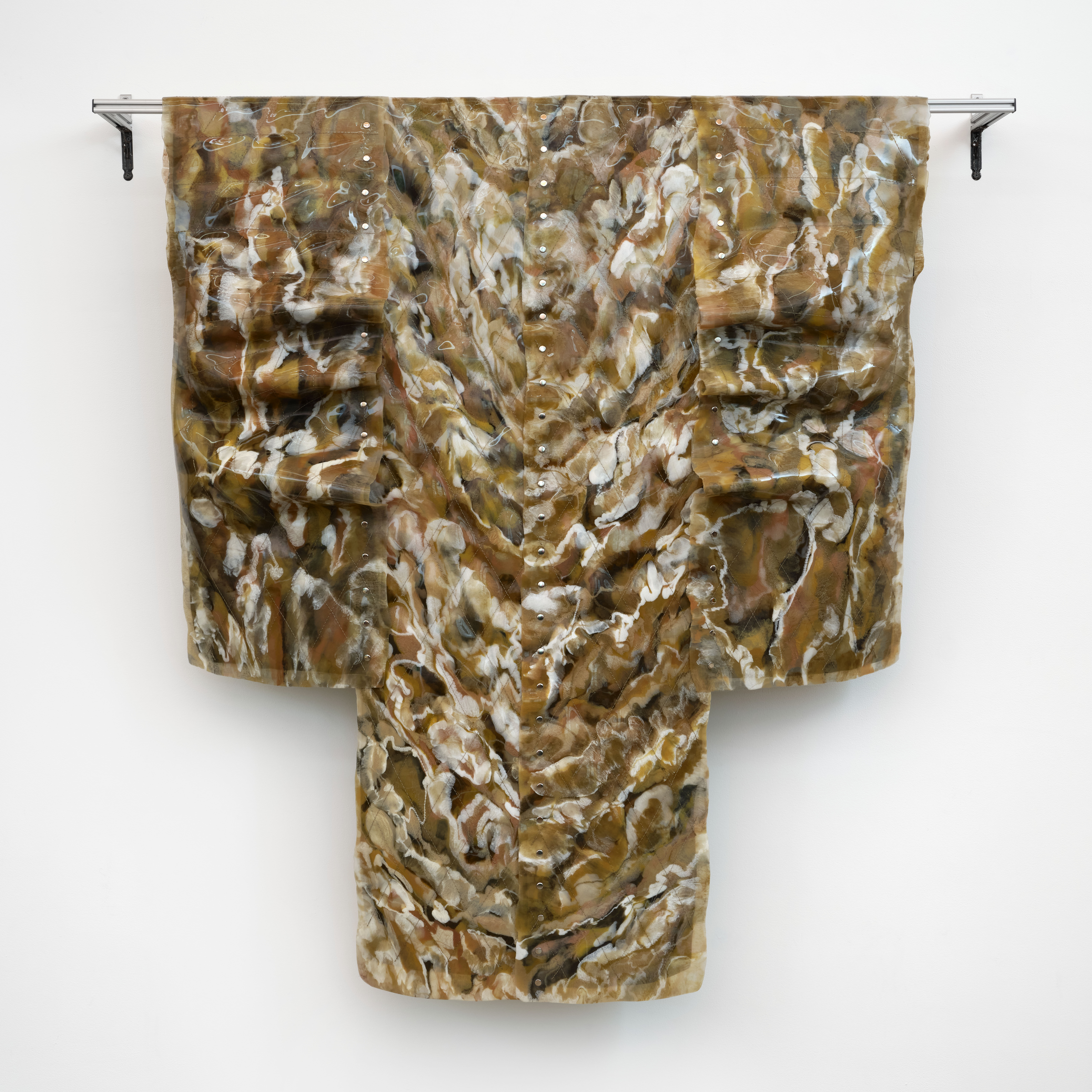 A wall-mounted sculpture made of poured silicone and other mixed materials, resembling a textured kimono hangs against a white wall. The surface features swirling patterns in earthy tones of brown, white, and beige.