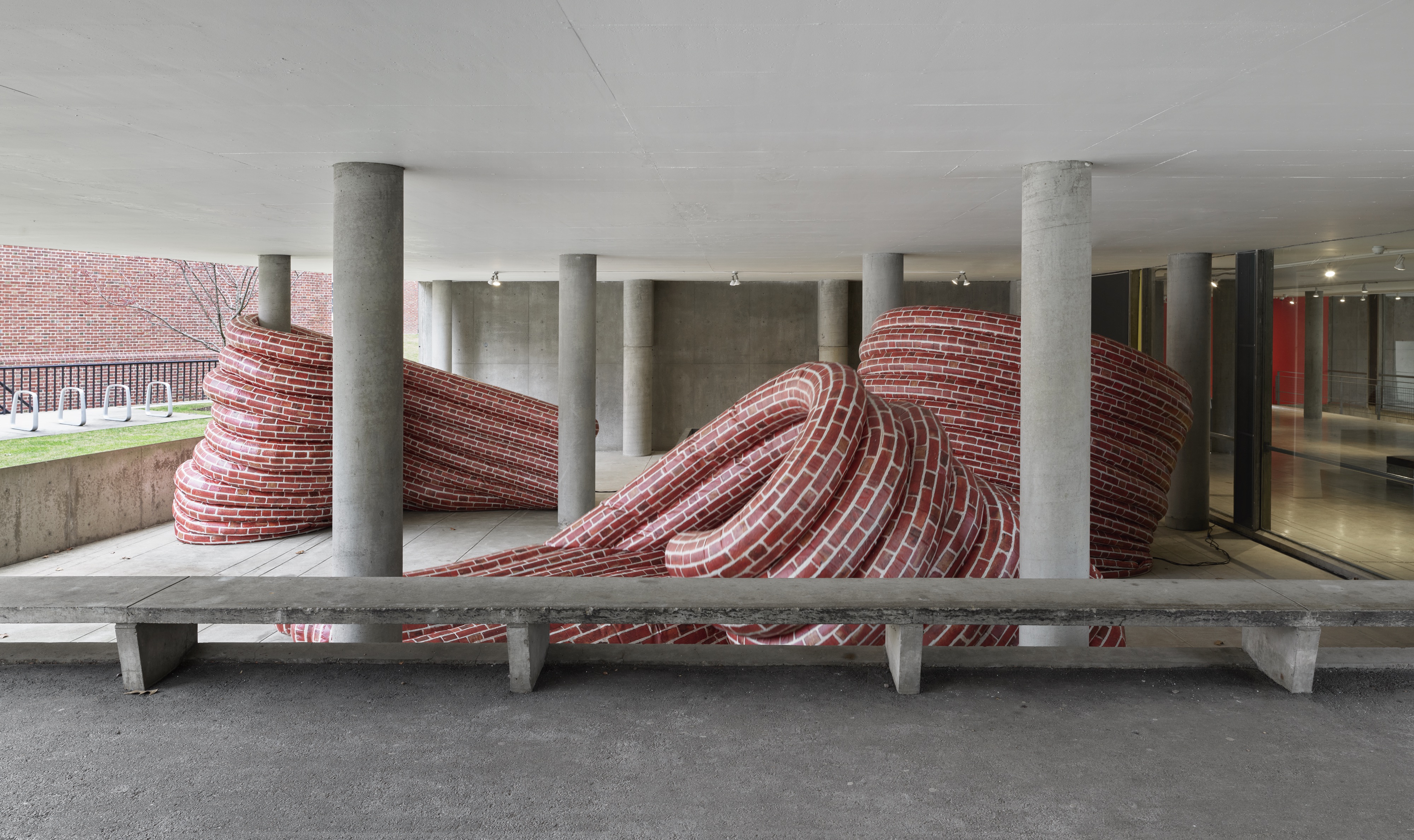 A view of an installation featuring curved inflatable walls patterned with red brick print situated at the entrance of a concrete building. The walls bend and twist into intricate forms, weaving between columns and a bench. The red color of the brick contrasts with the grey concrete.  