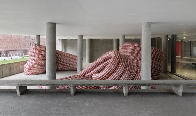 A view of an installation featuring curved inflatable walls patterned with red brick print situated at the entrance of a concrete building. The walls bend and twist into intricate forms, weaving between columns and a bench. The red color of the brick contrasts with the grey concrete.