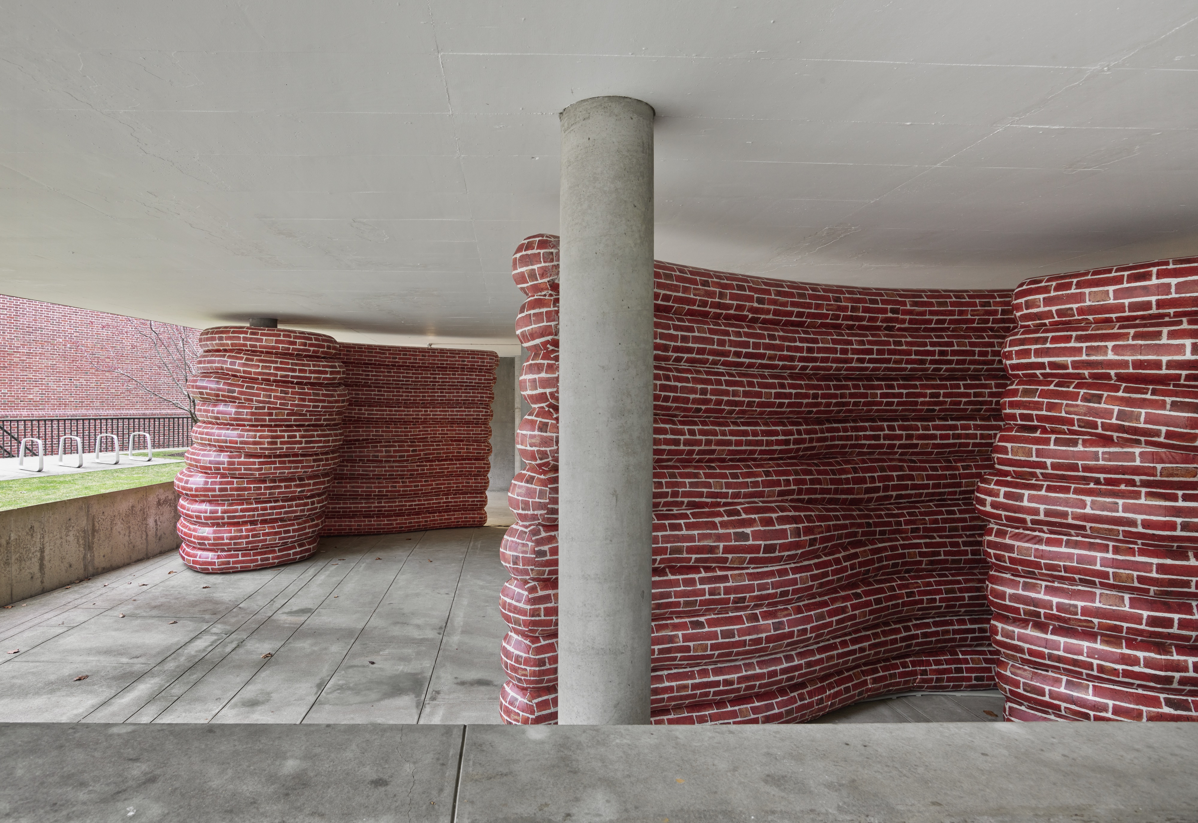 An installation featuring curved inflatable walls patterned with stacked red bricks wrap around concrete columns at the entrance of a building. The curvy red brick forms contrast with the grey concrete. 