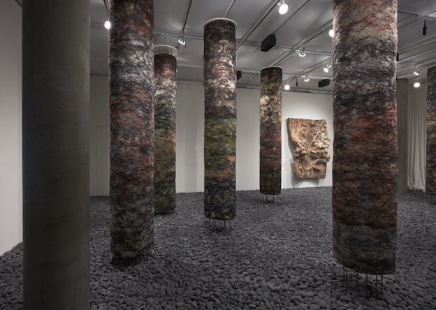A gallery filled with tall, cylindrical pillars wrapped in multicolored, wool-like organic material. The floor is covered in dark gray gravel. A large, abstract wall-mounted piece with folds and crevices is visible in the background.