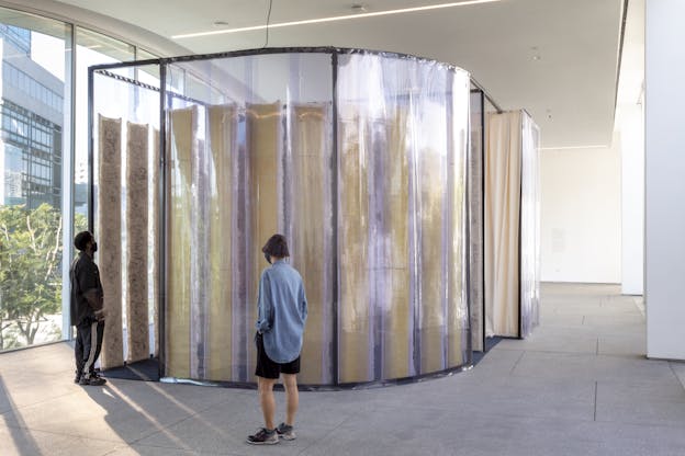 A curved installation made of translucent panels and felted wool layers stands in a brightly lit gallery space. Two viewers, one in a light blue shirt and another in dark clothing, observe the work closely. Large windows in the background reveal the surrounding cityscape and greenery.