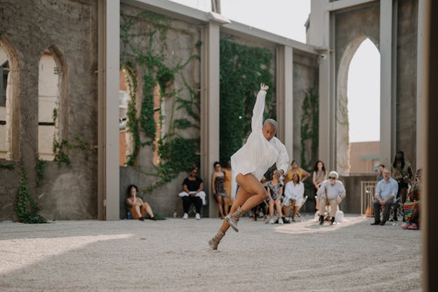 Jennifer Harge performs in an outdoor space with tall, ivy-covered walls and arched windows. She appears mid-step with one arm extended upward and her body leaning forward, and wears a white long-sleeved garment and lace-up boots. In the background, an audience watches attentively in the sunlit venue with gravel flooring.