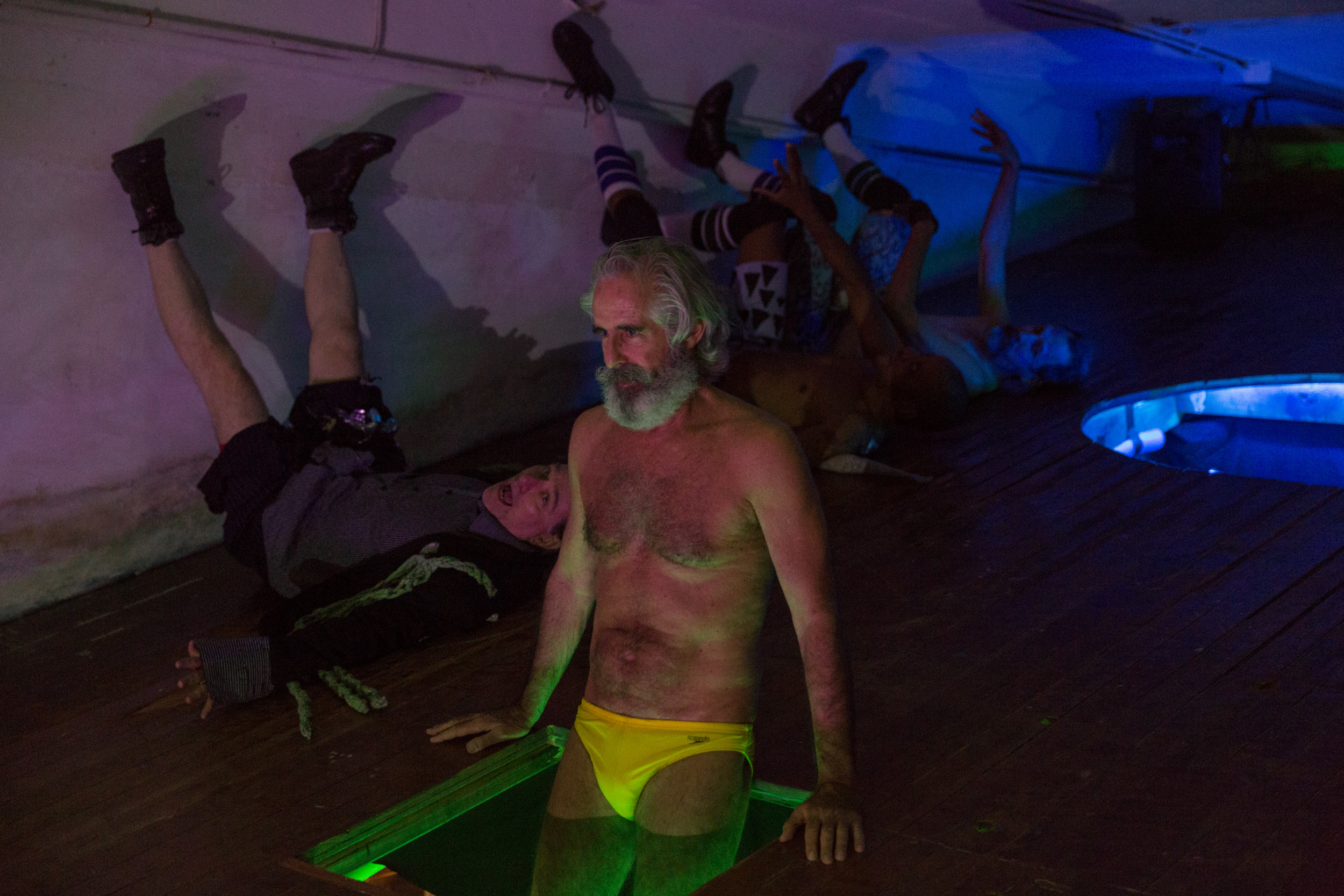 Philip Littell stands thigh-deep in a small rectangular pool of water on a wooden stage. The pool is illuminated green and he is wearing bright yellow briefs. Behind him, three figures recline on the floor with their legs raised against a wall, illuminated by purple and blue lighting.