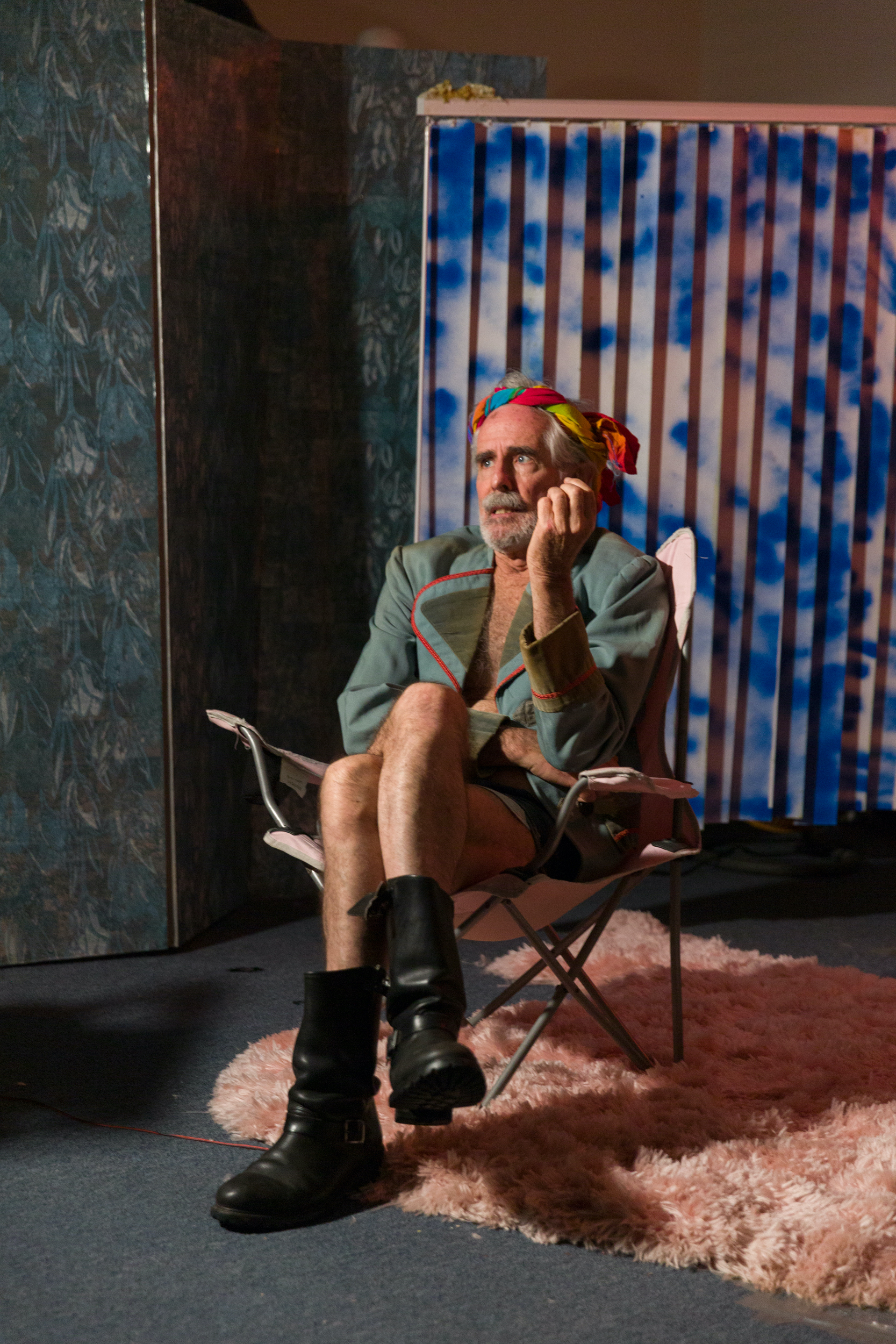 Philip Littell sits on a folding chair atop on a pink shag rug. He wears a rainbow headscarf, a green blazer with red trim, and black boots, with one leg crossed over the other. Behind him stands a patterned folding screen and a blue-and-pink accordion backdrop.