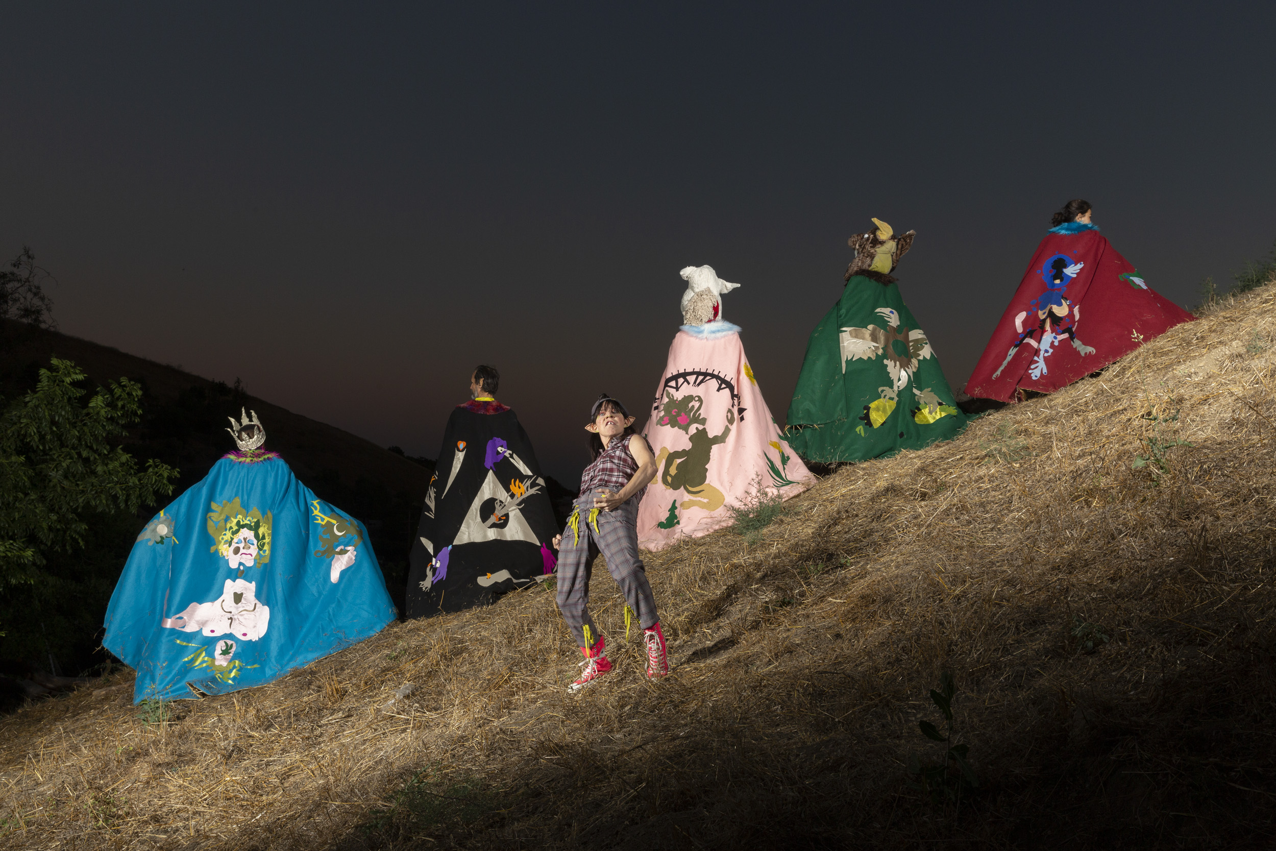 Six performers stand on a grassy hillside at dusk, their backs mostly turned toward the camera. From left to right, Paul Outlaw, Joe Seely, Michael Bonnabel, Zut Lorz, and Jacqueline Wright wear elaborate capes adorned with colorful, symbolic imagery. Jacqueline Wright, at the center, faces forward while striking a pose and wearing pointed ears, a plaid shirt and pants, and red sneakers.