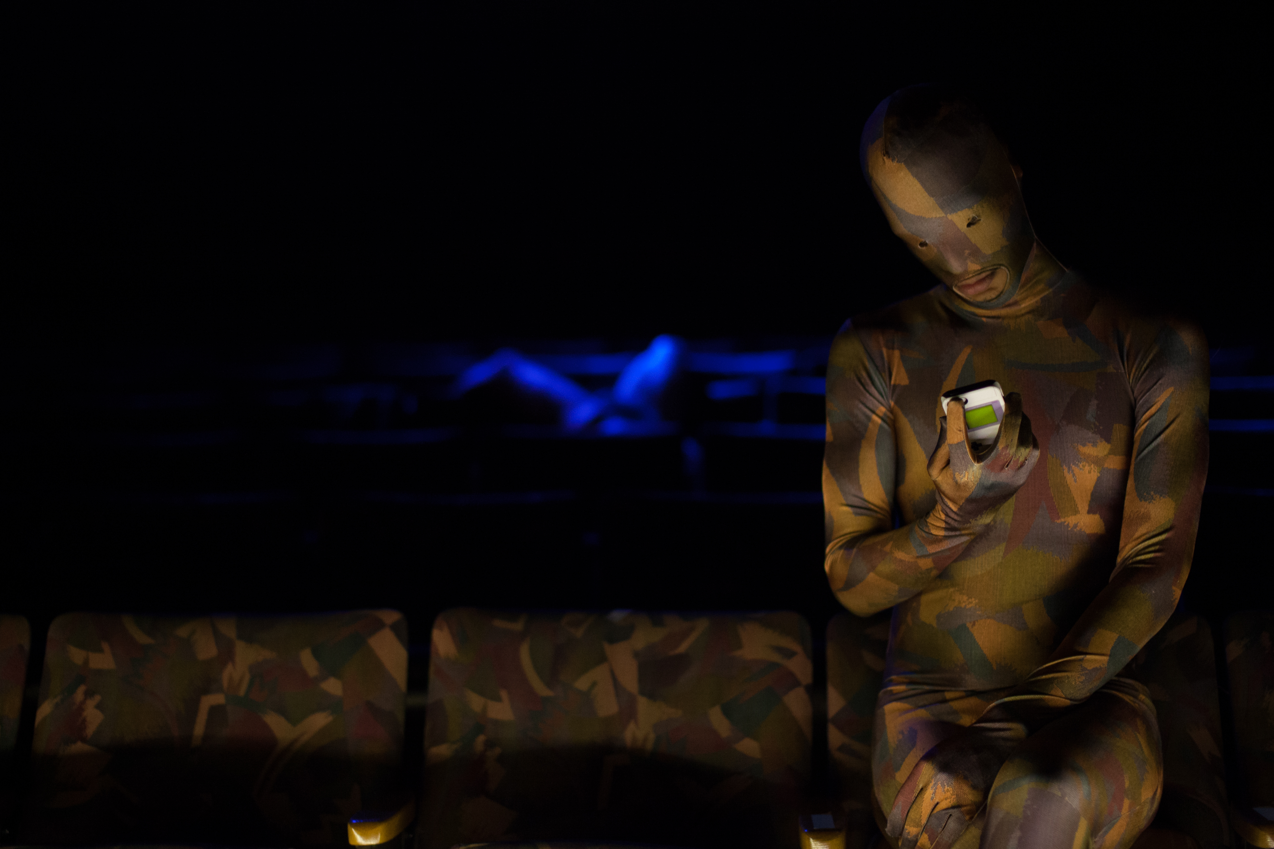 Bryatt Bryant and Philip Littell are pictured in a dimly lit theater space. Bryatt Bryant wears a full-body camouflage bodysuit and gazes at a mobile phone in his hand while seated in a patterned theater chair. Philip Littell, laying across several rows of seats in the background, is illuminated by blue light and is faintly visible.