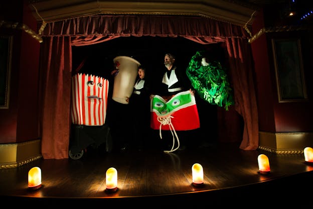 Drew Thataussie, Jasmine Orpilla, Philip Littell, and Chelsea Rector stand together on a small theatrical stage. They wear costumes and hold props of oversized food items, including a striped popcorn bucket, a soda cup, a leafy vegetable, and a pair of boxing shorts with a face. Stage lights illuminate the scene.